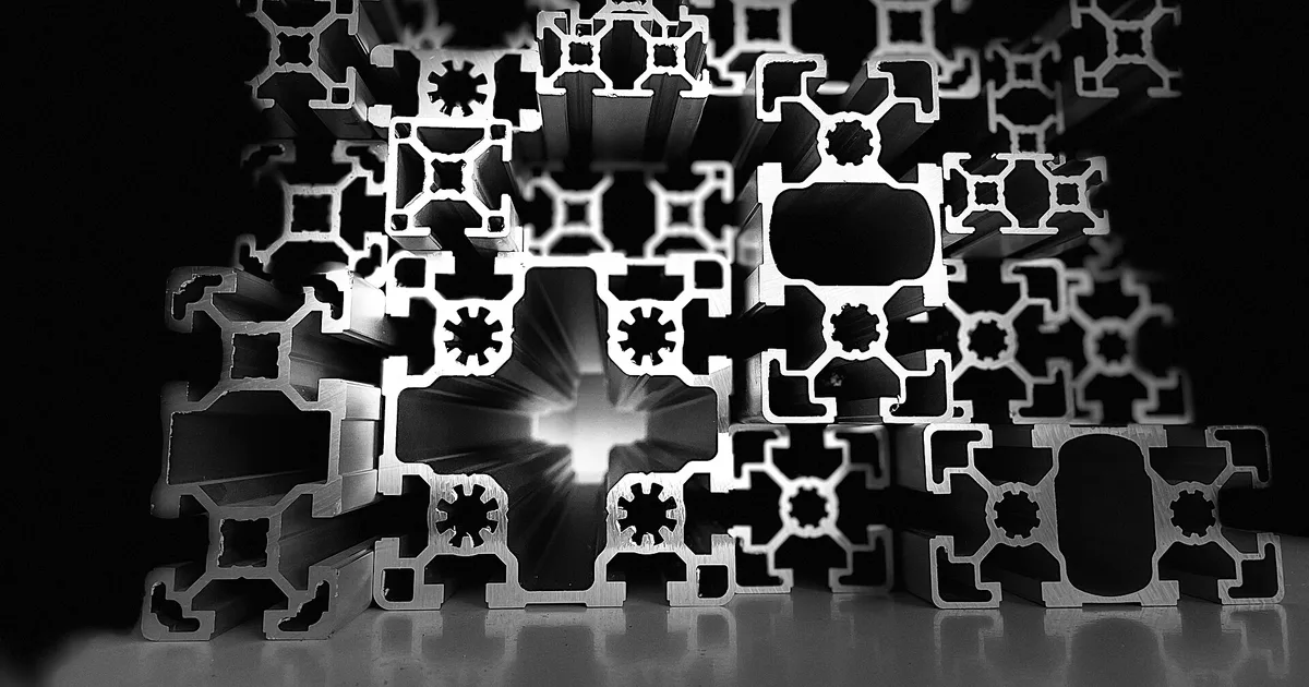 Black and white photo of stacked T-slot aluminium extrusion profiles seen end-on, showing hollow chambers and slots