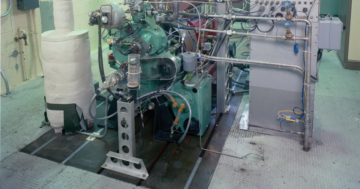 Engine test setup on a slotted steel floor plate in a test cell, with an insulated exhaust pipe and an instrumentation panel beside it