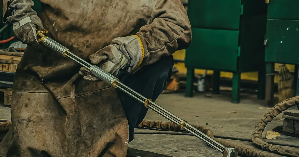 Worker in protective clothing holding a long gas torch near a steel workpiece in a fabrication shop, the equipment used to preheat thick plate before welding