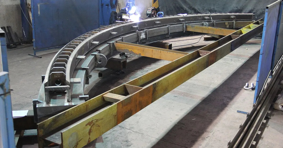 Large steel weldment held on a welding fixture in a fabrication shop, with a welder working at the far end of the frame